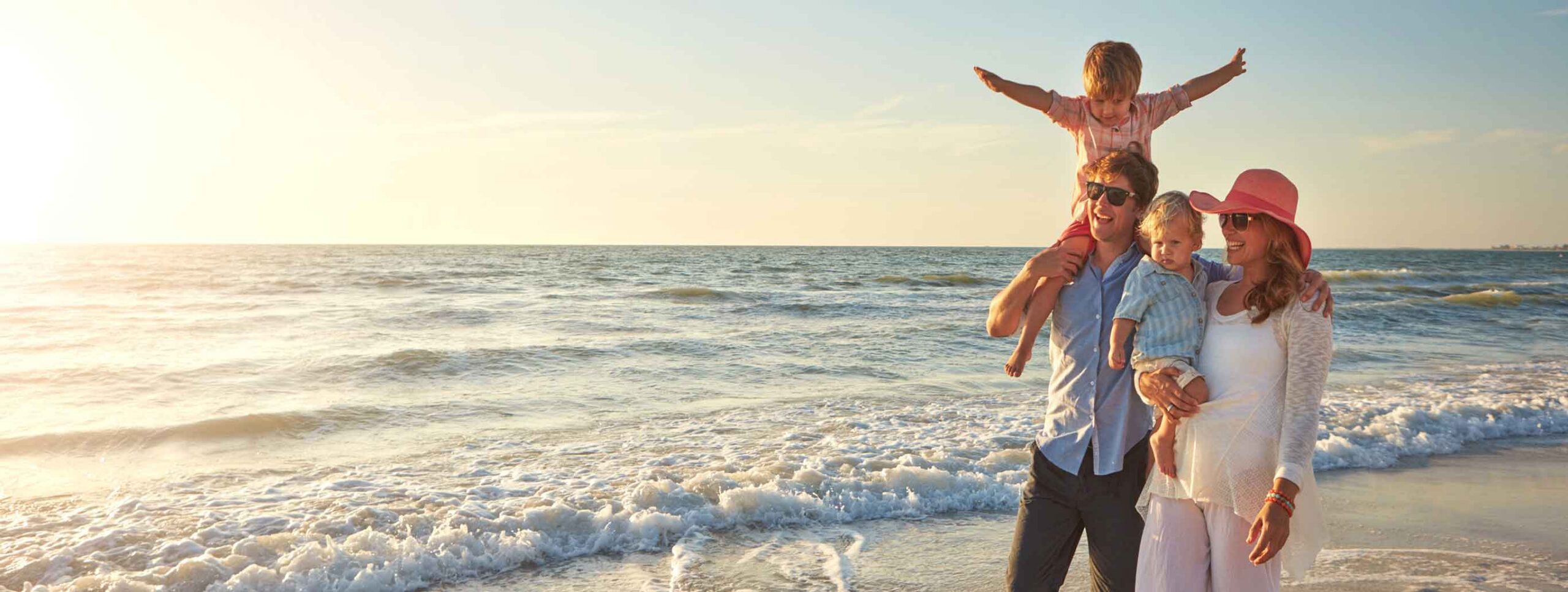 A family of four stands on a beach at sunset, smiling as they enjoy the ocean waves and sunshine—grateful for the easy start to their vacation thanks to Joe's Airport Parking.