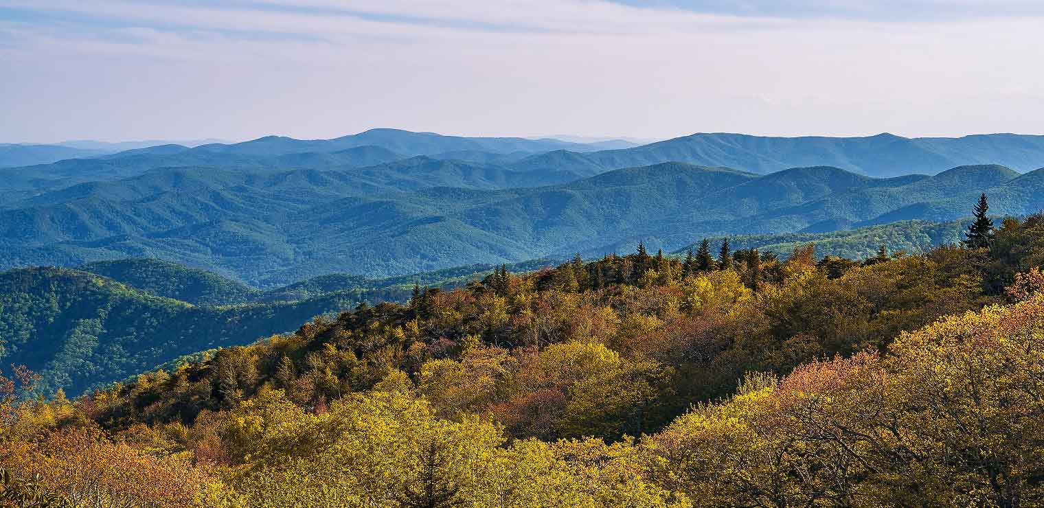 Rolling blue mountains stretch into the distance under a clear sky, with a foreground of dense autumn trees displaying shades of green, yellow, and orange—much like the vibrant scenery near Joe's Airport Parking.