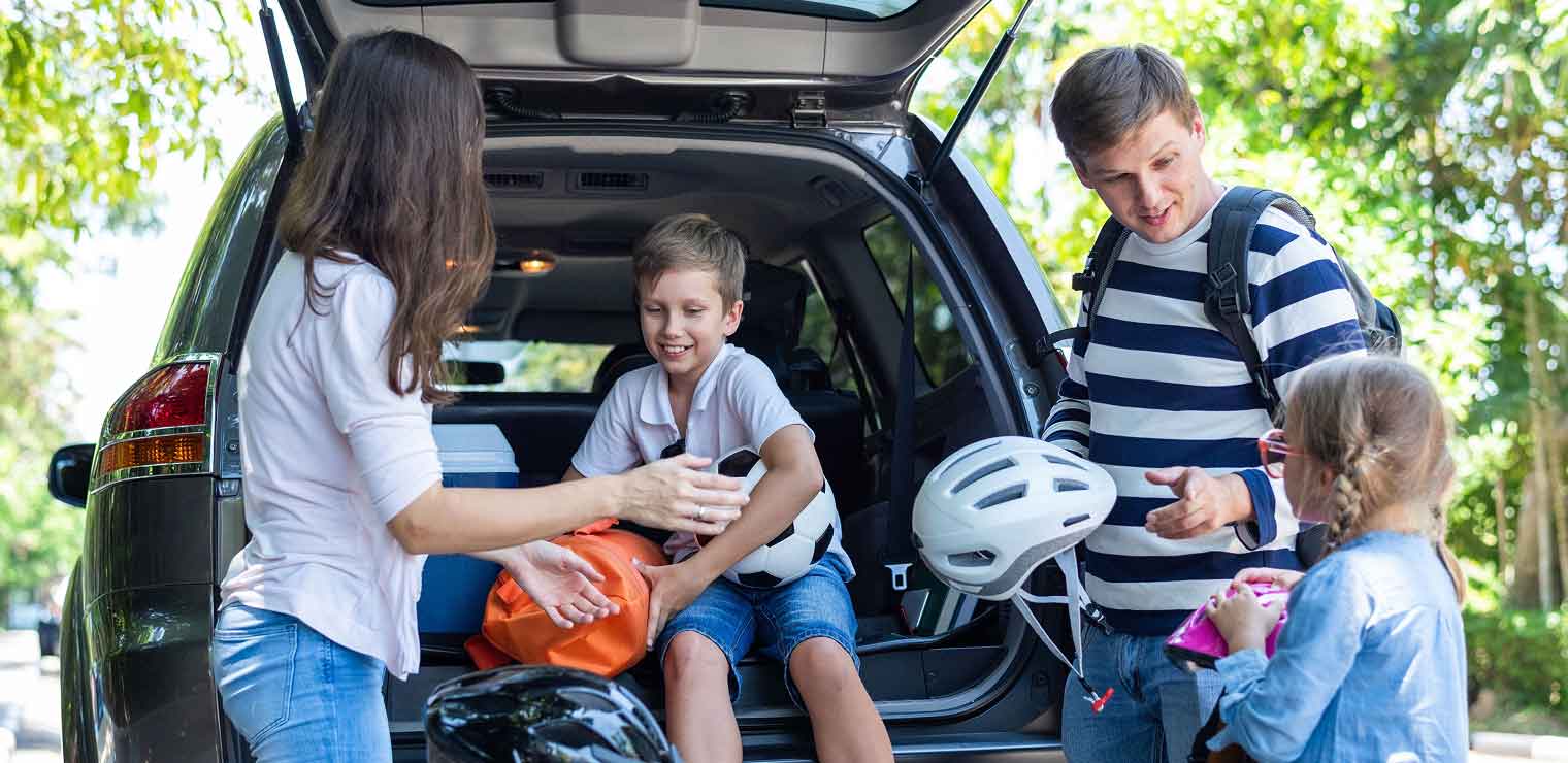 A family packs their SUV for a trip near Joe's Airport Parking. A woman hands a backpack to a smiling boy in the trunk, while a man with a white bike helmet chats with a girl holding her stuffed animal. Trees are visible in the background.