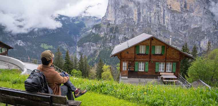 A person sits on a bench overlooking a wooden house, lush green grass, and dramatic mountains with clouds in the background—a peaceful scene far from the bustle of city life or the convenience of Joe’s Airport Parking.