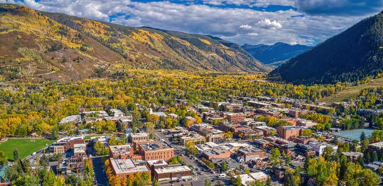 Aerial view of a small town surrounded by mountains and forests with fall foliage, under a partly cloudy sky, with Joe's Airport Parking conveniently located near the town center.
