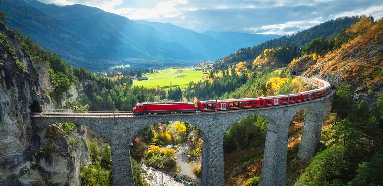 A red train crosses a tall stone bridge over a forested canyon, surrounded by green fields, mountains, and autumn-colored trees under a partly cloudy sky—not far from Joe's Airport Parking, where travelers begin their journeys amidst scenic landscapes.