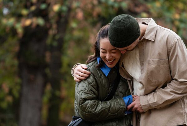 A couple stands outdoors, smiling and hugging each other closely, surrounded by green and brown foliage—perhaps savoring a moment together after a stress-free arrival thanks to Joe’s Airport Parking.