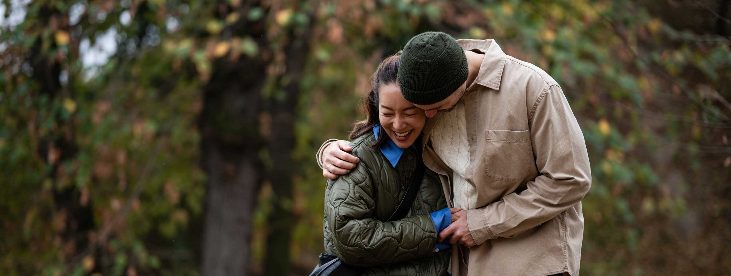 A couple stands outdoors, smiling and hugging each other closely, surrounded by green and brown foliage—perhaps savoring a moment together after a stress-free arrival thanks to Joe’s Airport Parking.