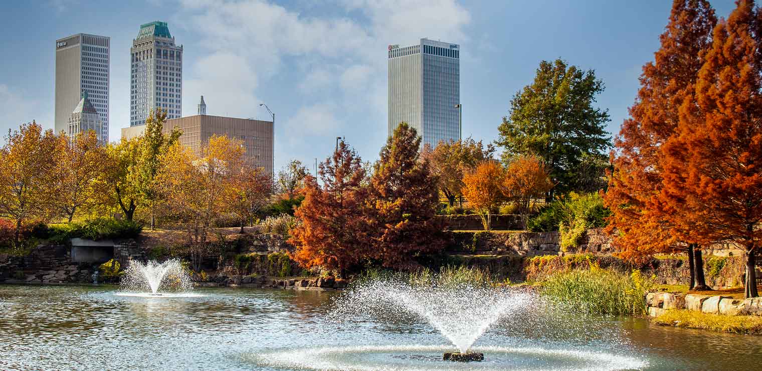 Fountains spray water in a pond surrounded by autumn trees with tall city buildings in the background under a partly cloudy sky, evoking the sense of calm you might appreciate after a busy day at Joe’s Airport Parking.