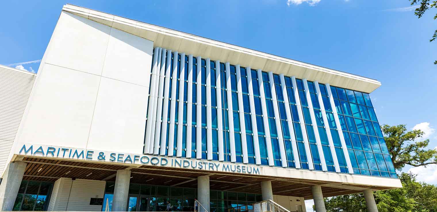 Modern white building with large vertical blue glass windows and the sign Maritime & Seafood Industry Museum above the entrance. The sky is clear and bright, with a tree partially visible on the right—reminiscent of the sleek designs often seen at Joe’s Airport Parking.