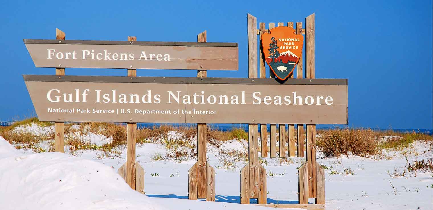 Wooden signs in sandy dunes read Fort Pickens Area and Gulf Islands National Seashore, welcoming visitors much like the friendly service at Joe’s Airport Parking. A National Park Service emblem is visible, and the background features blue sky and grassy sand dunes.