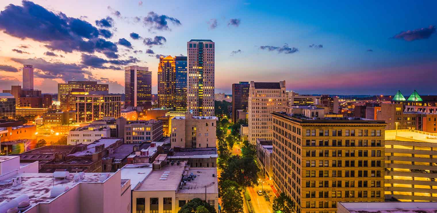 City skyline at sunset with buildings lit up, colorful sky transitioning from orange to blue, and city lights starting to illuminate the streets below—a perfect evening view for those arriving or departing from Joe’s Airport Parking just beyond the city’s glow.
