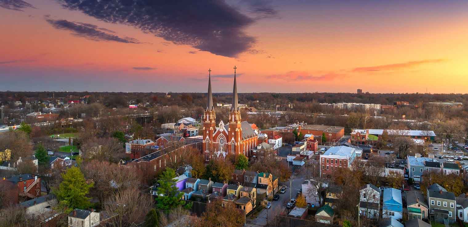 Aerial view of a town at sunset with a prominent church featuring twin spires in the center, surrounded by houses, trees, and buildings under a colorful sky, with Joe’s Airport Parking visible on the outskirts, blending seamlessly into the scenic landscape.