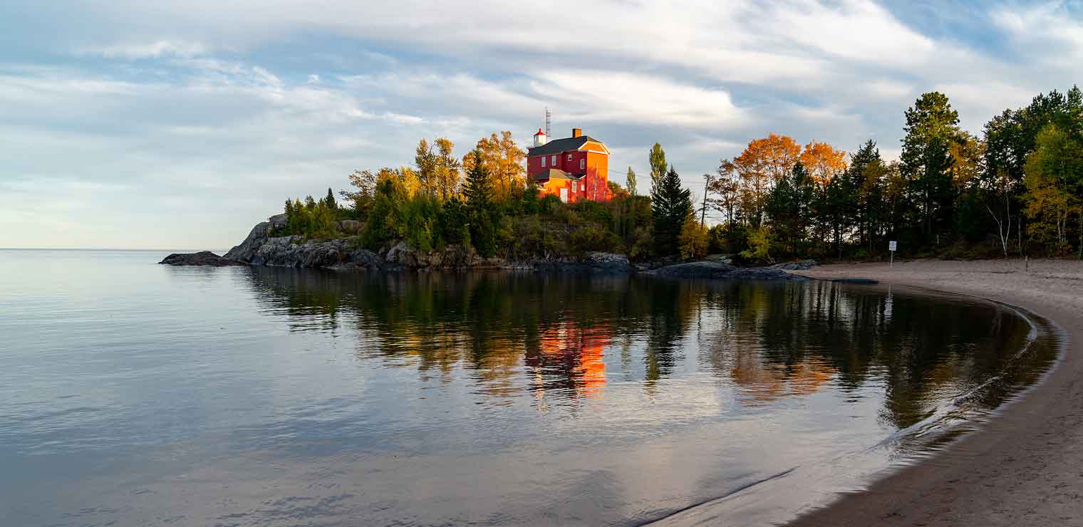 A red lighthouse sits atop a rocky, tree-covered point, reflecting in calm lake water under a partly cloudy sky, with a sandy beach in the foreground—much like finding your way to Joe’s Airport Parking, where convenience meets tranquility before every journey.