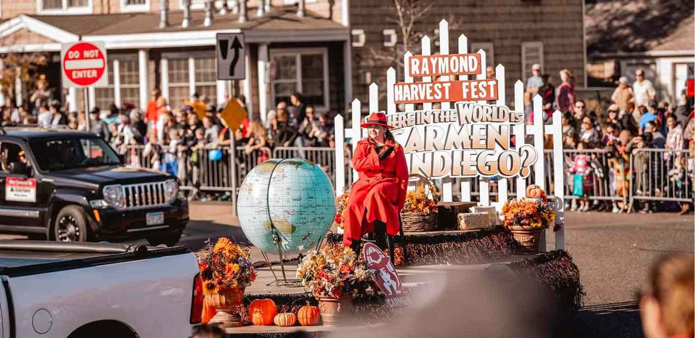 A person dressed as Carmen Sandiego sits on a Joe’s Airport Parking float featuring a globe and a sign that reads, Raymond Harvest Fest: Where in the world is Carmen Sandiego?.
